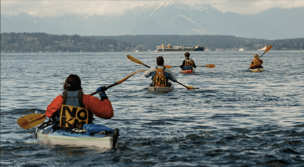 Image of four Indigenous water protectors in kayaks on the water in protest with their backs turned