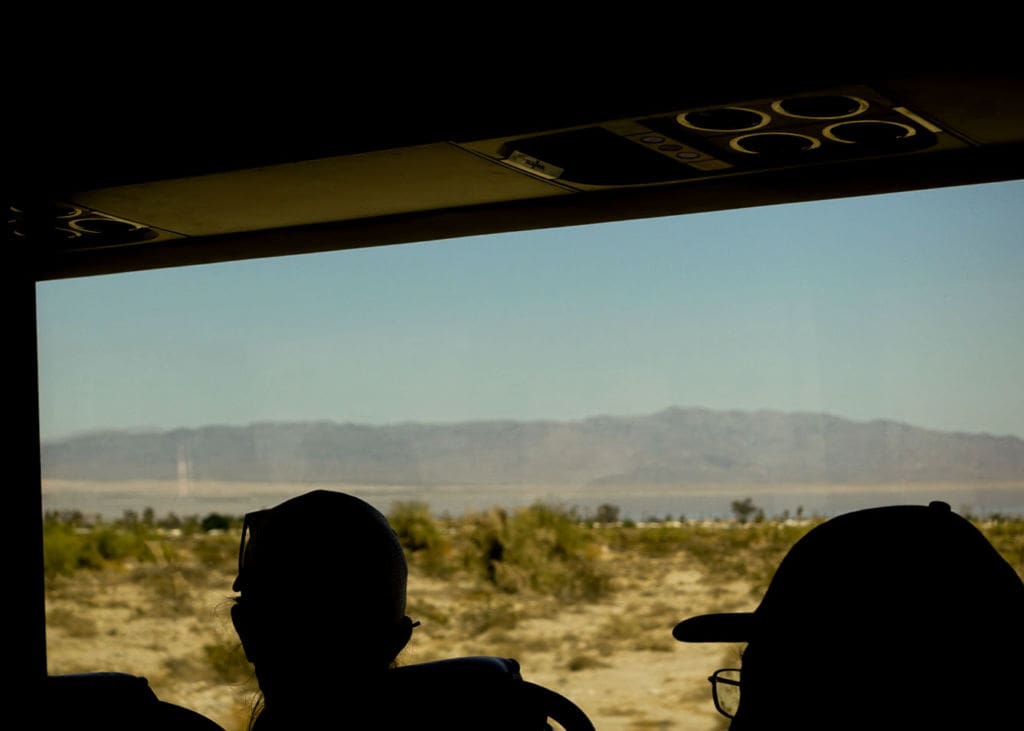 Photo of silhouettes of two people in front of a bus window with yellow-green brush in the landscape.