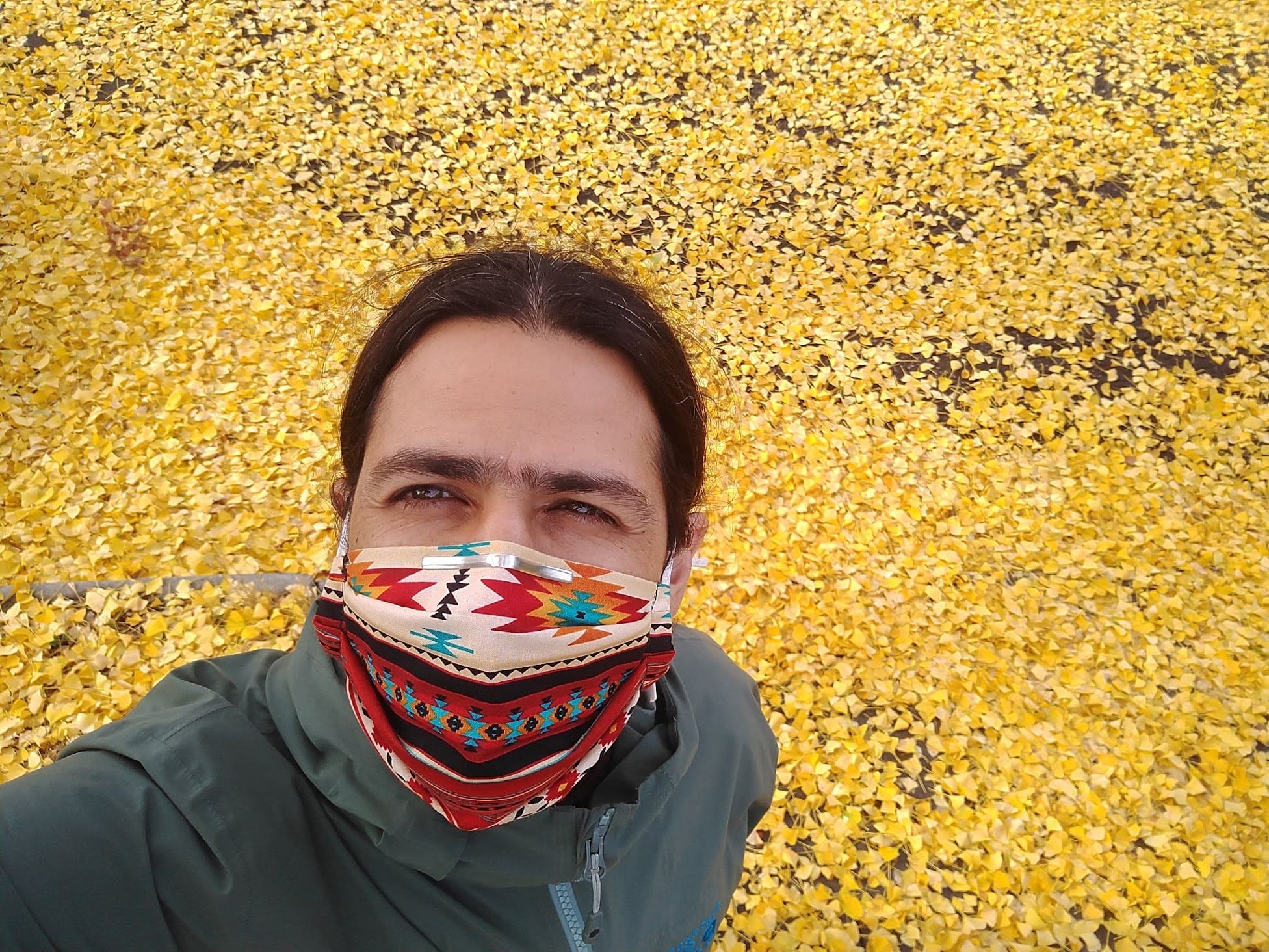 High-angle selfie of José wearing a face cover with colorful Native American designs. The background is filled with thousands of yellow maple leaves that cover the floor. He was taking a walk in Berkeley, CA, during the fall and right after it rained.
