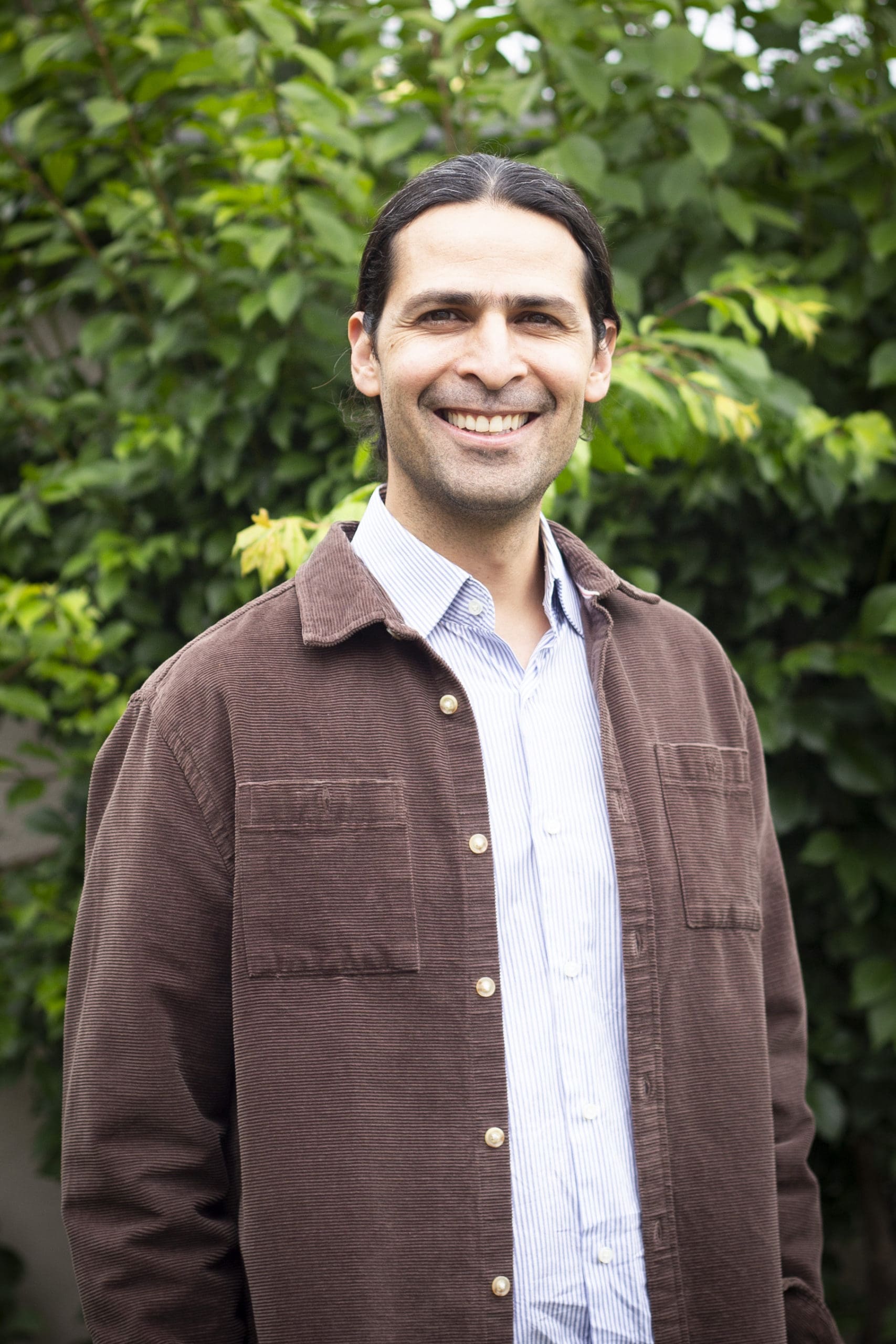 Headshot of José Aranda smiling with a brown jacket on standing in front of greenery outside