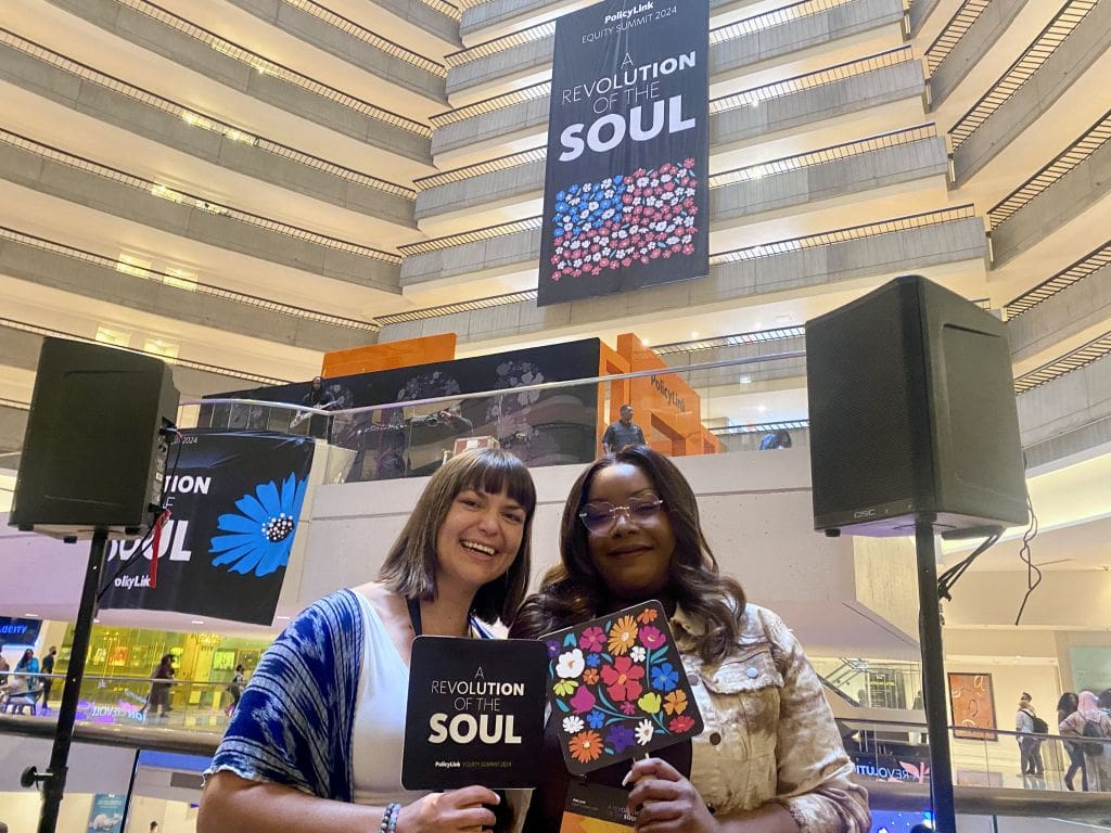 Sarah and Zakiyaa pose at the 2024 PolicyLink Equity Summit in front of a sign with text "A Revolution of the Soul" and a depiction of the U.S. flag made up with red, white, and blue flowers.