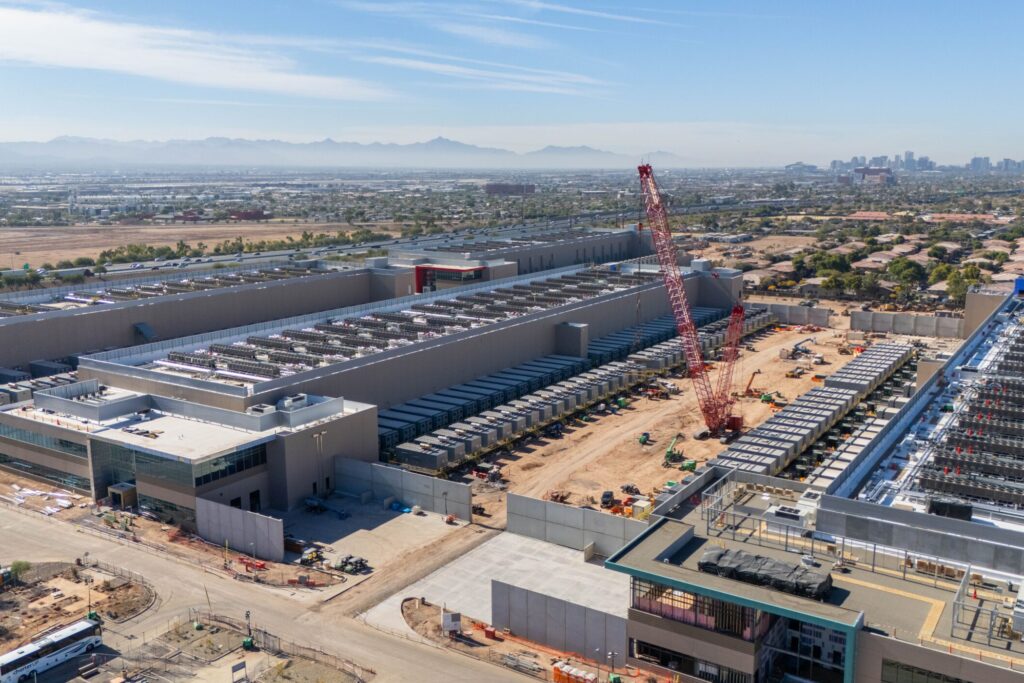 An aerial shot shows the construction site of a sprawling industrial facility. The scene is set during daytime under a clear blue sky. Several large buildings are already in place, surrounded by construction equipment and red cranes that are actively working.