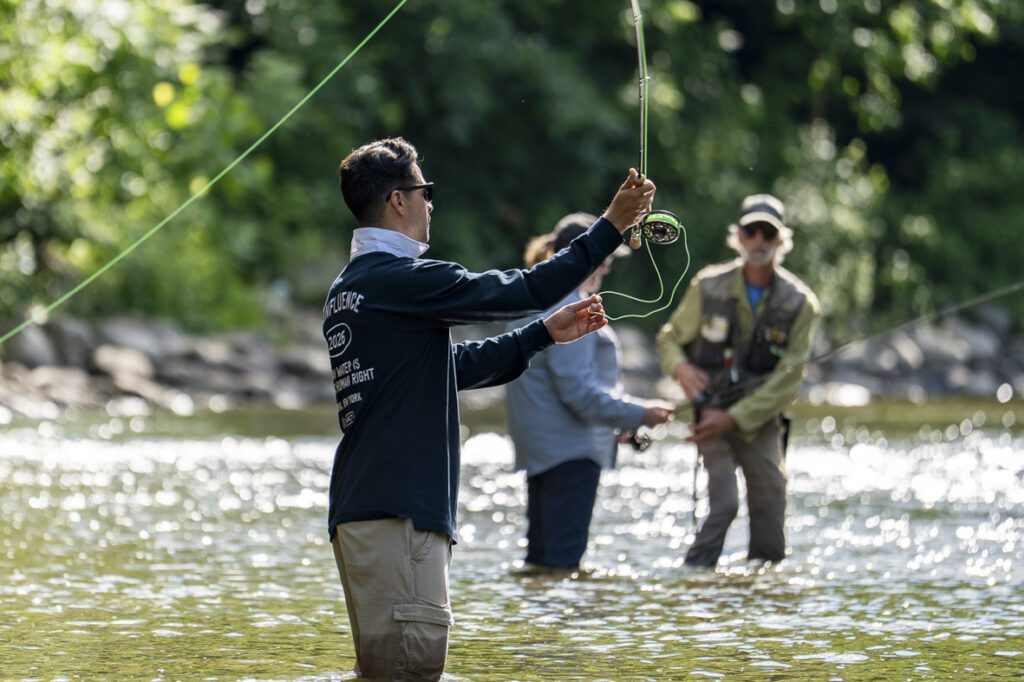 A person viewed from the side wears a dark navy blue long-sleeve rugby-style shirt with a white collar. White graphic text is printed on the back and sleeve, including the phrase "CLEAN WATER IS A HUMAN RIGHT." The person wears khaki pants and holds a green fly-fishing line over a sparkling, sunlit river. In the blurred background, another person in a gray shirt stands in the water.