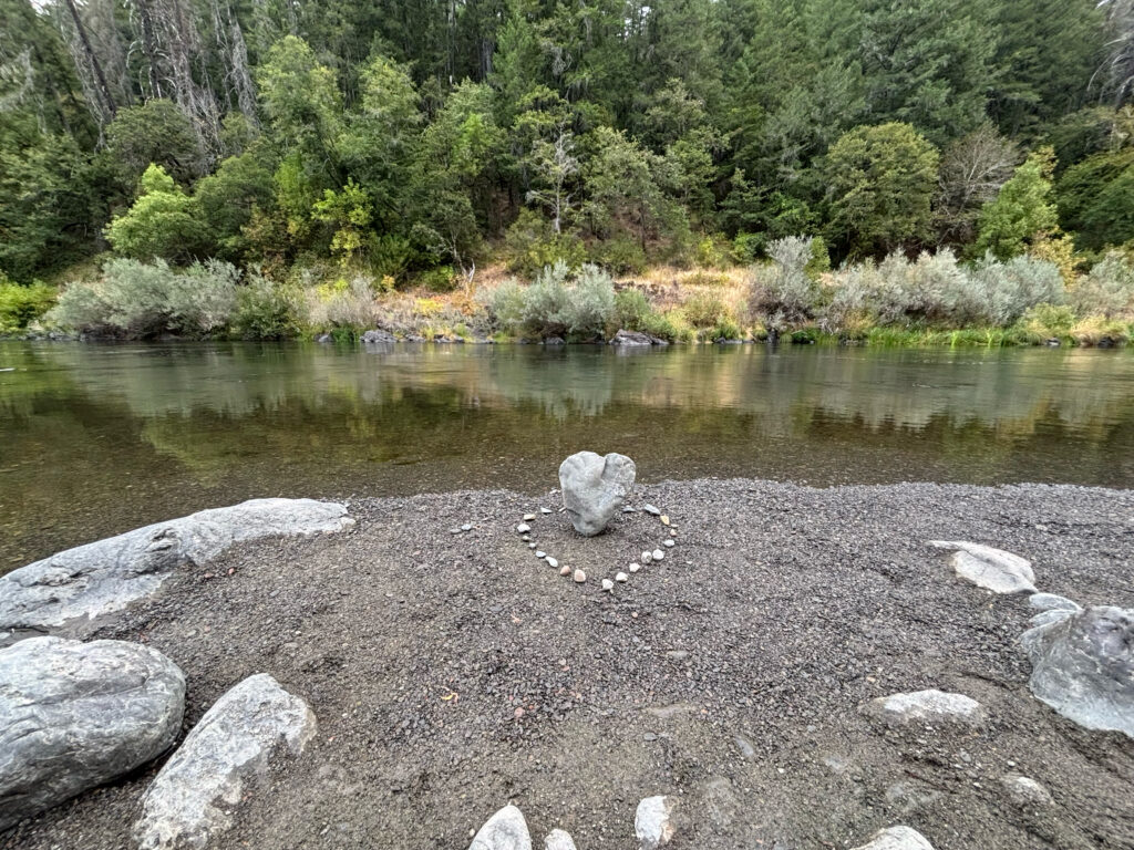 A heart-shaped rock surrounded by pebbles in the shape of a heart on the shore of a river.